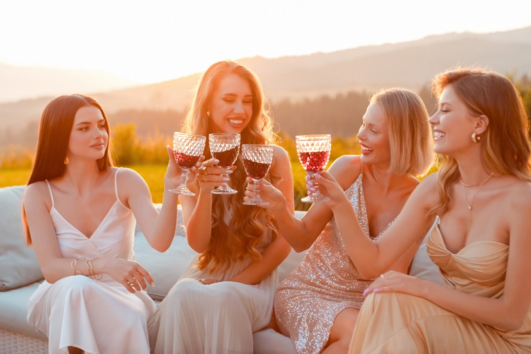 Female friends wearing elegant dresses sitting on sofa outdoors and toasting with red wine at sunset. Young women during bachelorette party.