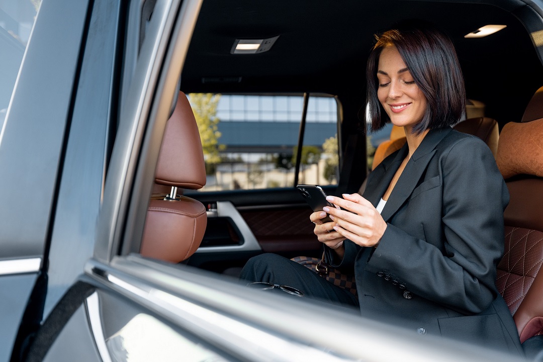 Portrait of an elegant business lady in black formal wear sitting with phone on backseat of luxury SUV vehicle. Concept of business trips and transportation